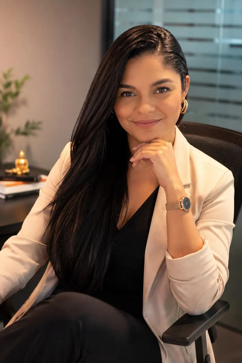 Sheila Batista no escritório, de blazer claro, com a mão no queixo