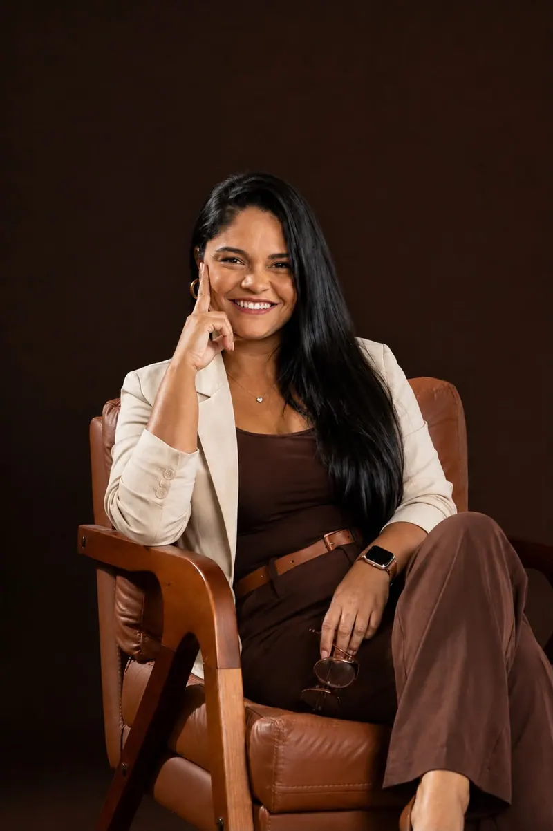 Sheila Batista sentada na poltrona de couro, com a mão no rosto