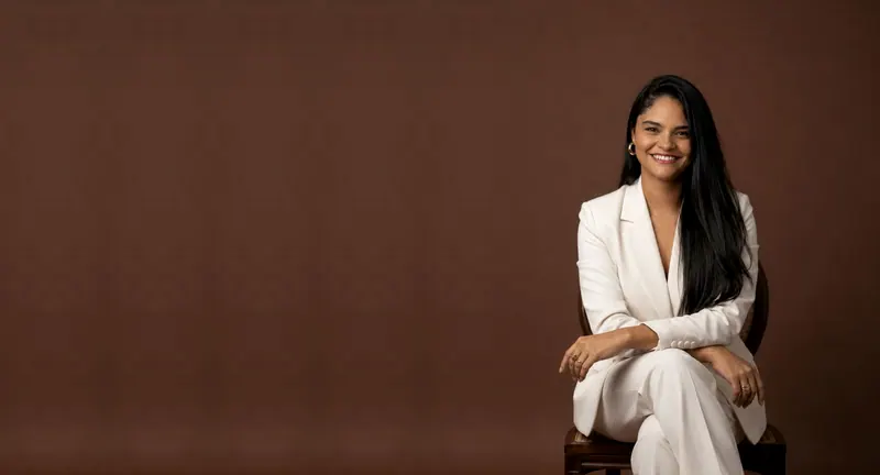 Sheila Batista de terno branco, sorrindo, sentada numa cadeira de madeira no estúdio de fundo marrom